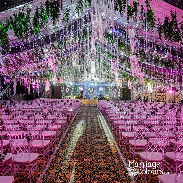 A grand wedding venue with pink chairs, a patterned aisle, and a ceiling adorned with greenery and lights.