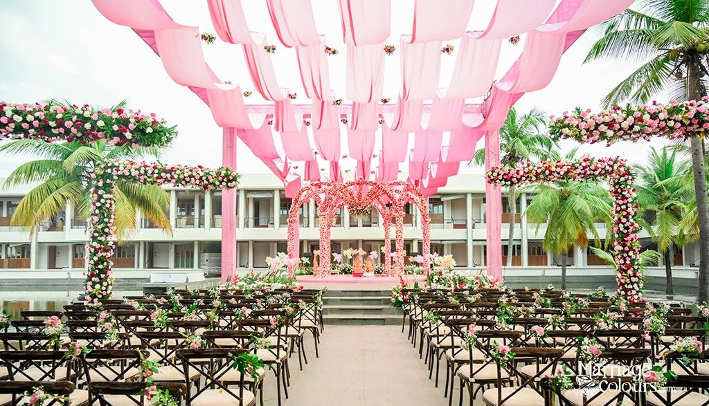 An outdoor wedding venue with a pink canopy, floral decorations, and a stage set against a backdrop of palm trees and a building.