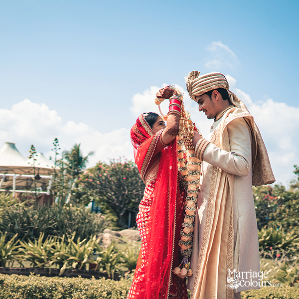 A bride in a red lehenga and groom in a cream sherwani exchange floral garlands during an outdoor wedding ceremony under a bright blue sky.