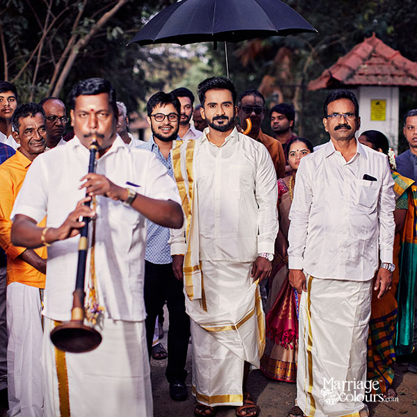 A groom in traditional white and gold attire walking in a procession, accompanied by a musician playing a wind instrument, with guests and an umbrella overhead.