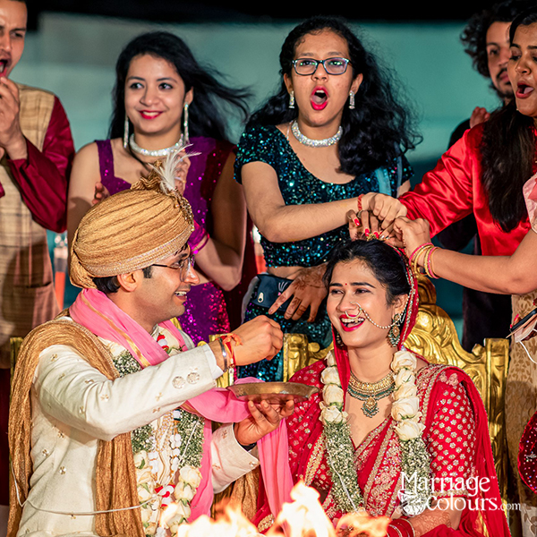 A bride and groom performing a traditional Hindu wedding ritual, with the groom feeding the bride a sweet dish while guests cheer and celebrate.