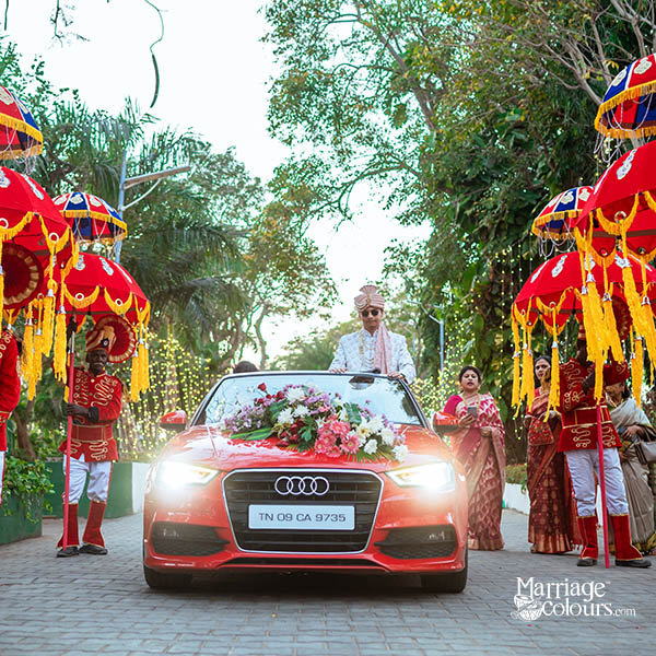 A red Audi car decorated with flowers, serving as the wedding car, with a groom standing on the roof, surrounded by attendants holding traditional red and yellow umbrellas.