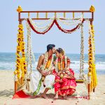 A bride and groom sharing an intimate moment during a traditional South Indian beach wedding, seated under a floral mandap decorated with marigold and jasmine garlands.