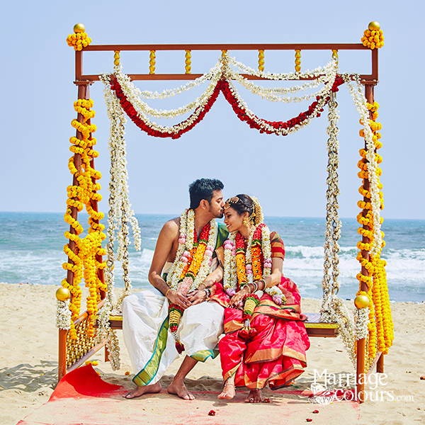 A bride and groom sharing an intimate moment during a traditional South Indian beach wedding, seated under a floral mandap decorated with marigold and jasmine garlands.
