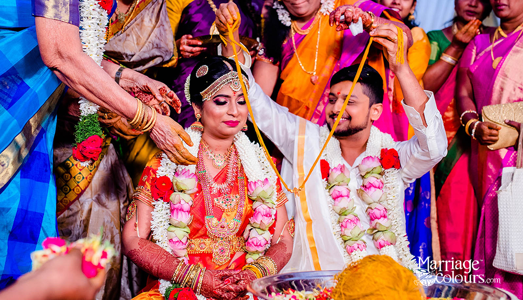 A bride and groom during a traditional South Indian wedding ceremony, exchanging sacred threads as family members bless them, surrounded by colorful sarees and floral garlands.