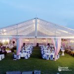 Large clear-span event tent set up on a lawn at twilight, featuring a ceiling of fairy lights, pink and blue draped fabric columns, round tables with white linens and blue sashes, and chandeliers inside.