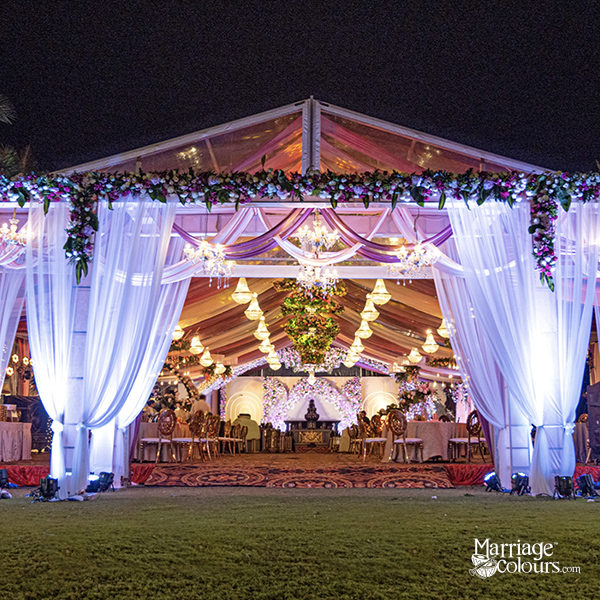Large clear-span event tent set up on a lawn at twilight, featuring a ceiling of fairy lights, pink and blue draped fabric columns, round tables with white linens and blue sashes, and chandeliers inside.
