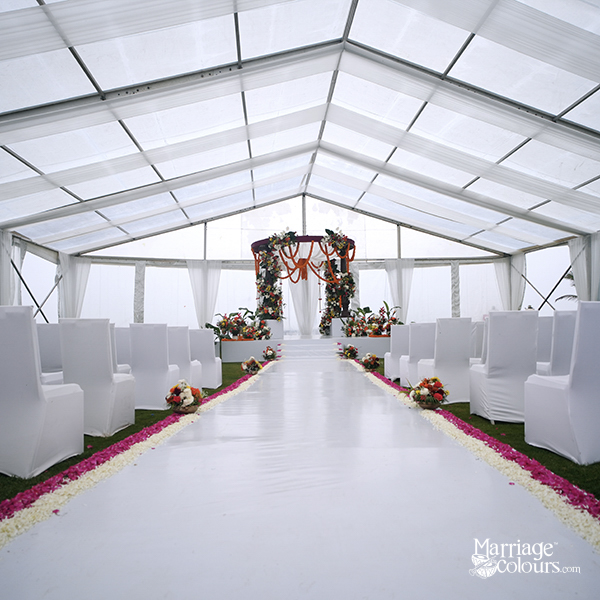 Interior view of a clear-span white event tent set up for a wedding ceremony, featuring a white aisle runner lined with pink and white flower petals, rows of white-draped chairs, and a floral mandap or altar at the far end.