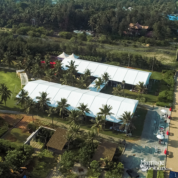 Aerial daytime view of a large beachside event venue featuring two long white clear-span tents surrounded by palm trees, with a sandy beach and ocean visible to the right and lush greenery in the background.