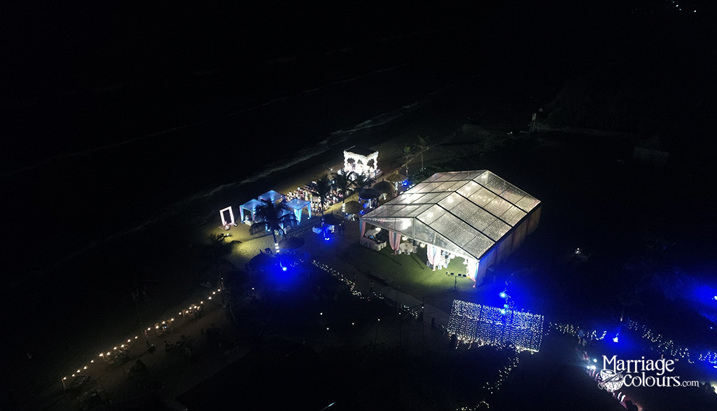 Aerial night view of a beachside event setup with illuminated tents, string lights, and a dance floor near the shoreline. Blue accent lighting and warm white decor create a festive atmosphere.