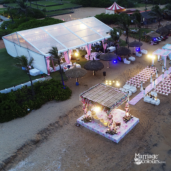 Aerial twilight view of a beach wedding setup featuring a decorated mandap (wedding altar) on the sand, rows of white guest chairs, a pink aisle runner, and a large illuminated white event tent in the background with thatched umbrellas and ambient lighting.